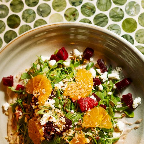 A colorful salad with greens, orange slices, red vegetables, and cheese crumbles in a bowl on a patterned table.