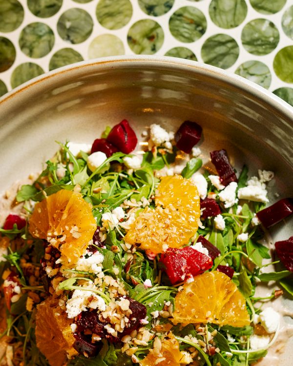 A colorful salad with greens, orange slices, red vegetables, and cheese crumbles in a bowl on a patterned table.