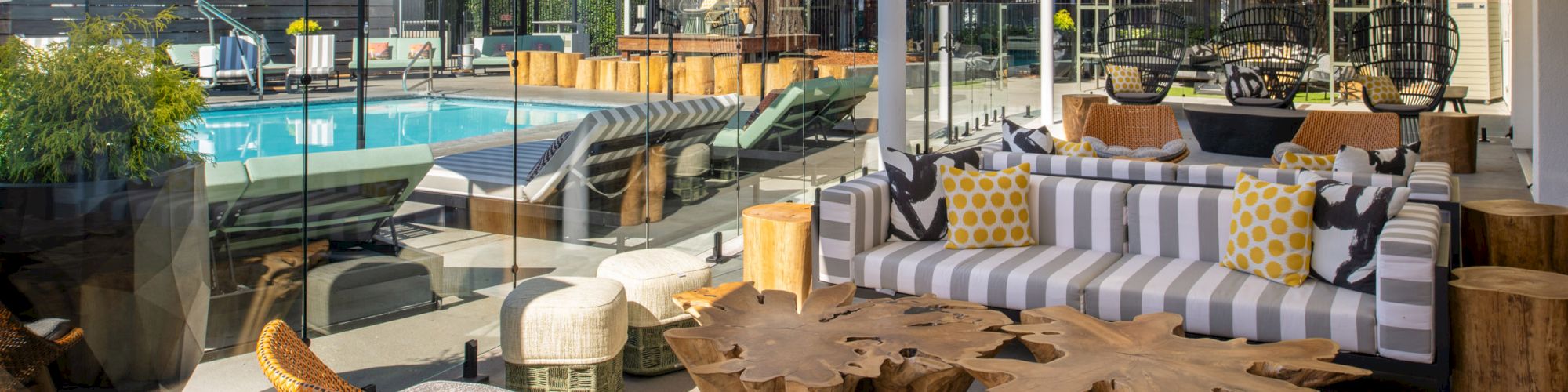 Outdoor lounge with cushioned seating, wood tables, and rattan chairs near a poolside area, featuring umbrellas and modern decor, during the daytime.