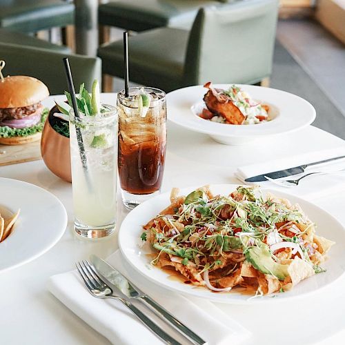 Various dishes including nachos, a burger, and drinks are on a white table in a restaurant setting, with modern green chairs and a bright atmosphere.