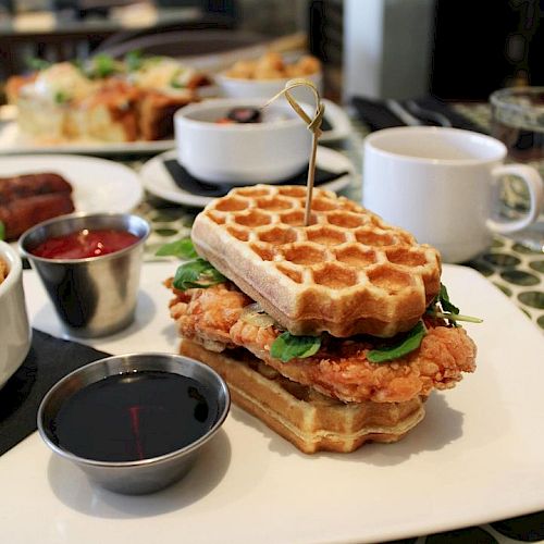 This image shows a waffle sandwich with fried chicken, spinach, and other fillings on a white plate. Accompaniments include syrup and ketchup.