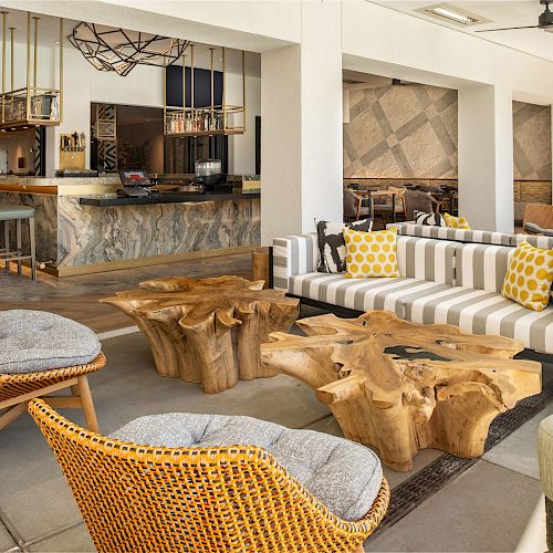 A stylish indoor bar lounge area with striped couches, wooden tables, wicker chairs, and modern decor, creating a cozy and inviting atmosphere.