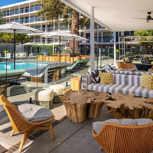 The image shows an outdoor seating area with wooden furniture, a lounge area, and a swimming pool in the background, under a shaded patio.