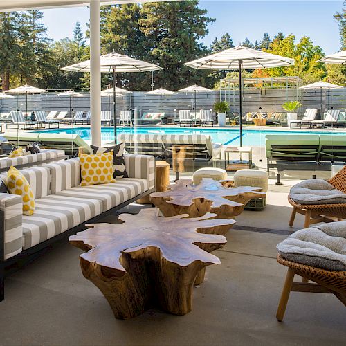 The image shows a modern poolside lounge area with striped sofas, a unique wooden table, cushioned chairs, and umbrellas beside a swimming pool.