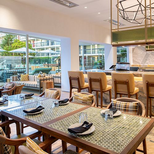 A modern dining area with tables and chairs, a bar with stools, and large windows showing an outdoor seating area by a pool.