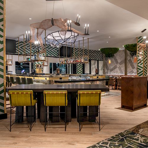 The image shows a modern bar and restaurant with colorful decor, featuring green and yellow stools, geometric wall patterns, and hanging plants.