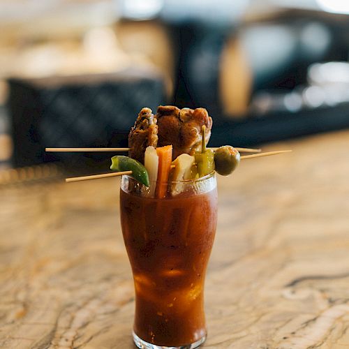 A glass of Bloody Mary garnished with celery, olives, bacon, green beans, and peppers, placed on a marble counter with a blurred background.