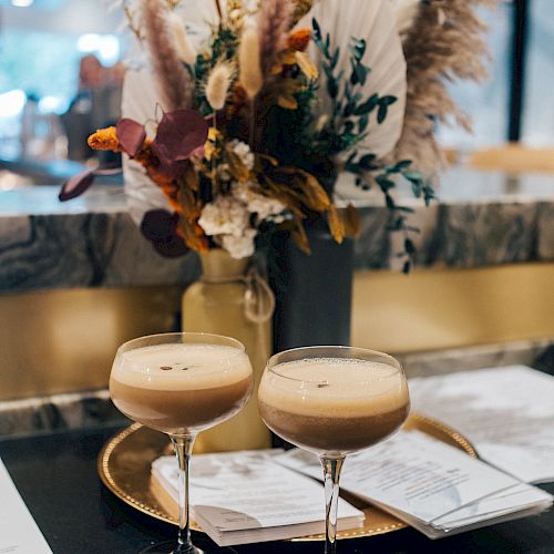 Two cocktail glasses with drinks sit on a table near a vase of floral arrangements, accompanied by some papers or menus in the background.