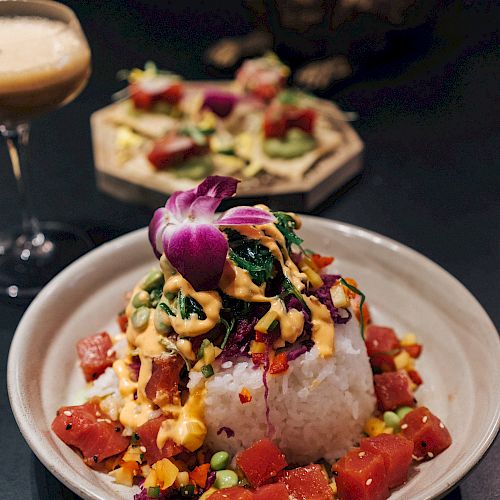 A bowl of rice topped with various colorful ingredients, possibly a poke bowl, accompanied by a drink and appetizer in the background.