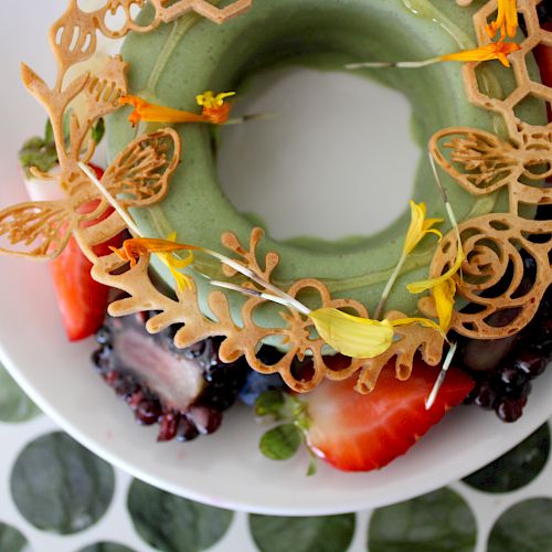 A decorative dessert with green icing, fresh berries, and intricate edible toppers shaped like bees and hexagons on a white plate.