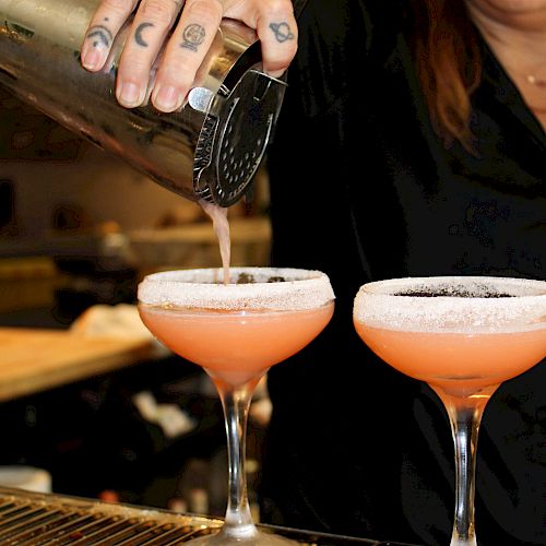 A bartender is pouring a pink cocktail into two glasses with sugar-rimmed edges, using a strainer and shaker.