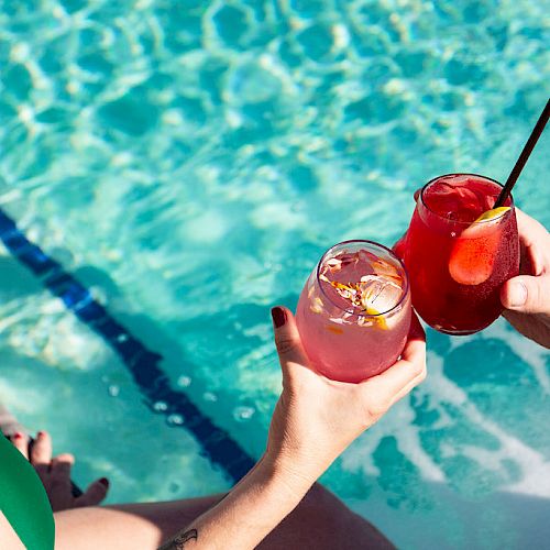 Two people are holding colorful drinks near a pool, their hands and part of their bodies visible. Both wear swimsuits.