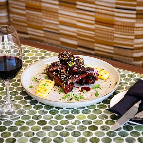 A plate of food and a glass of red wine on a patterned table, with utensils and a napkin on the side, are shown in this image.