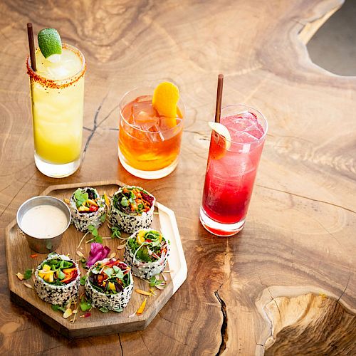 Three colorful drinks and a platter of sushi rolls with a dipping sauce are placed on a wooden table.