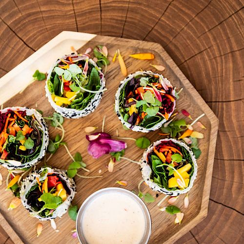 This image shows a wooden tray with six colorful vegetable rolls and a small dish of sauce in the center.