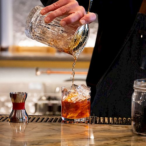 A bartender pours a cocktail from a mixing glass into an ice-filled glass on a bar counter, with a jigger nearby and a marble surface.