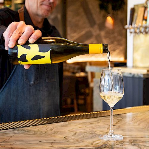 A person in an apron is pouring white wine from a bottle into a wine glass on a marble counter.