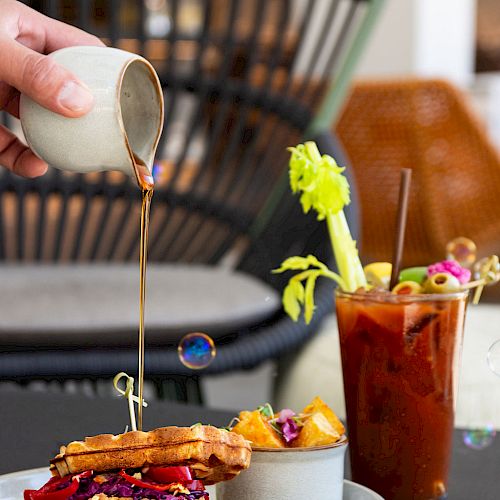 A hand pours syrup onto a stack of waffles with fried chicken, accompanied by a side of potatoes and a glass of garnished tomato juice.