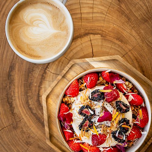 A latte in a cup beside a bowl of granola with strawberries, dried flowers, and various toppings on a wooden surface.