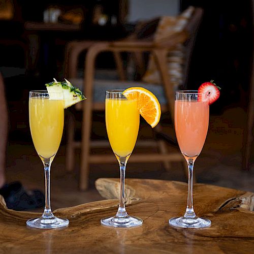 Three colorful cocktails garnished with pineapple, orange, and strawberry slices, placed on a wooden table in a cozy setting.