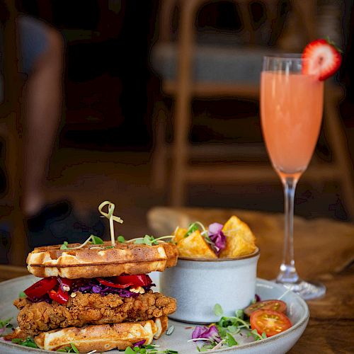 The image shows a plate with a fried chicken sandwich between waffles, a side of potatoes, and a garnished drink with a strawberry slice on a wooden table.