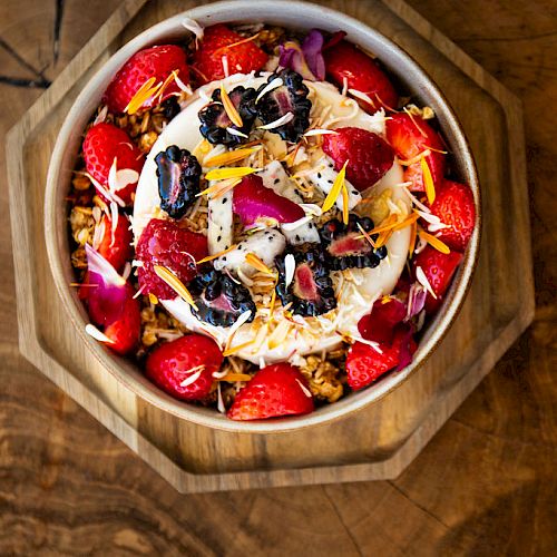 The image depicts a bowl of granola topped with yogurt, strawberries, blackberries, nuts, and colorful edible flowers, placed on a wooden surface.