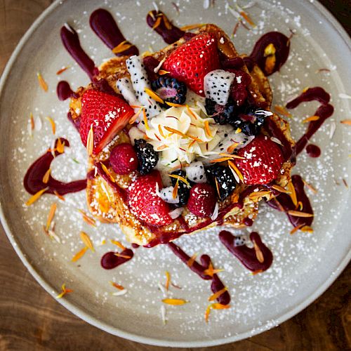 A plate of French toast topped with strawberries, dragon fruit, berries, and powdered sugar, garnished with syrup and a few edible flowers.