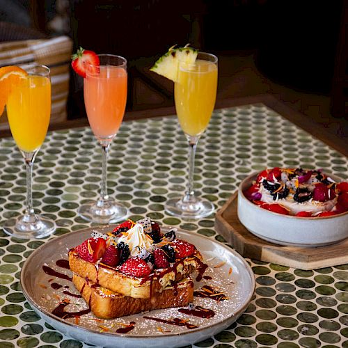 Three colorful cocktails and two delicious desserts, one on a plate and one in a bowl, on a patterned table.