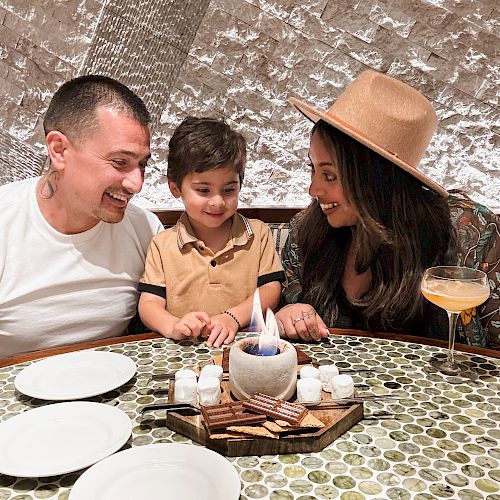 A family of three sits at a table with a small flame in the center, surrounded by plates. The child looks at the flame while the parents smile.