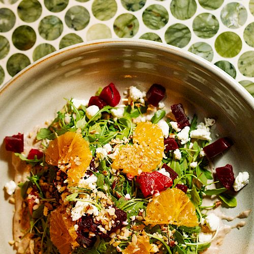 A salad in a bowl with orange slices, red beet pieces, greens, and crumbled cheese is on a green polka-dotted table surface.