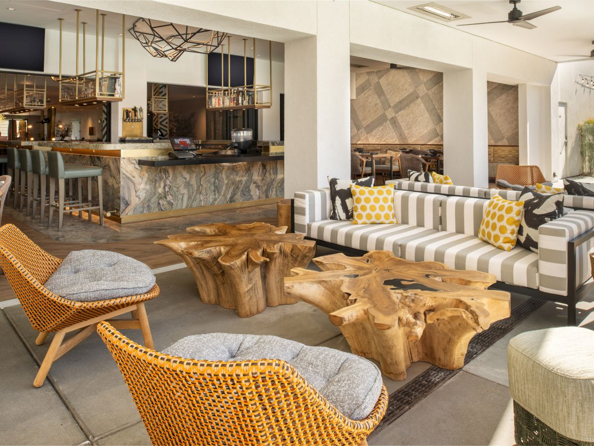 This image shows a stylish indoor seating area with wooden furniture, wicker chairs, and modern decor, complemented by a bar in the background.