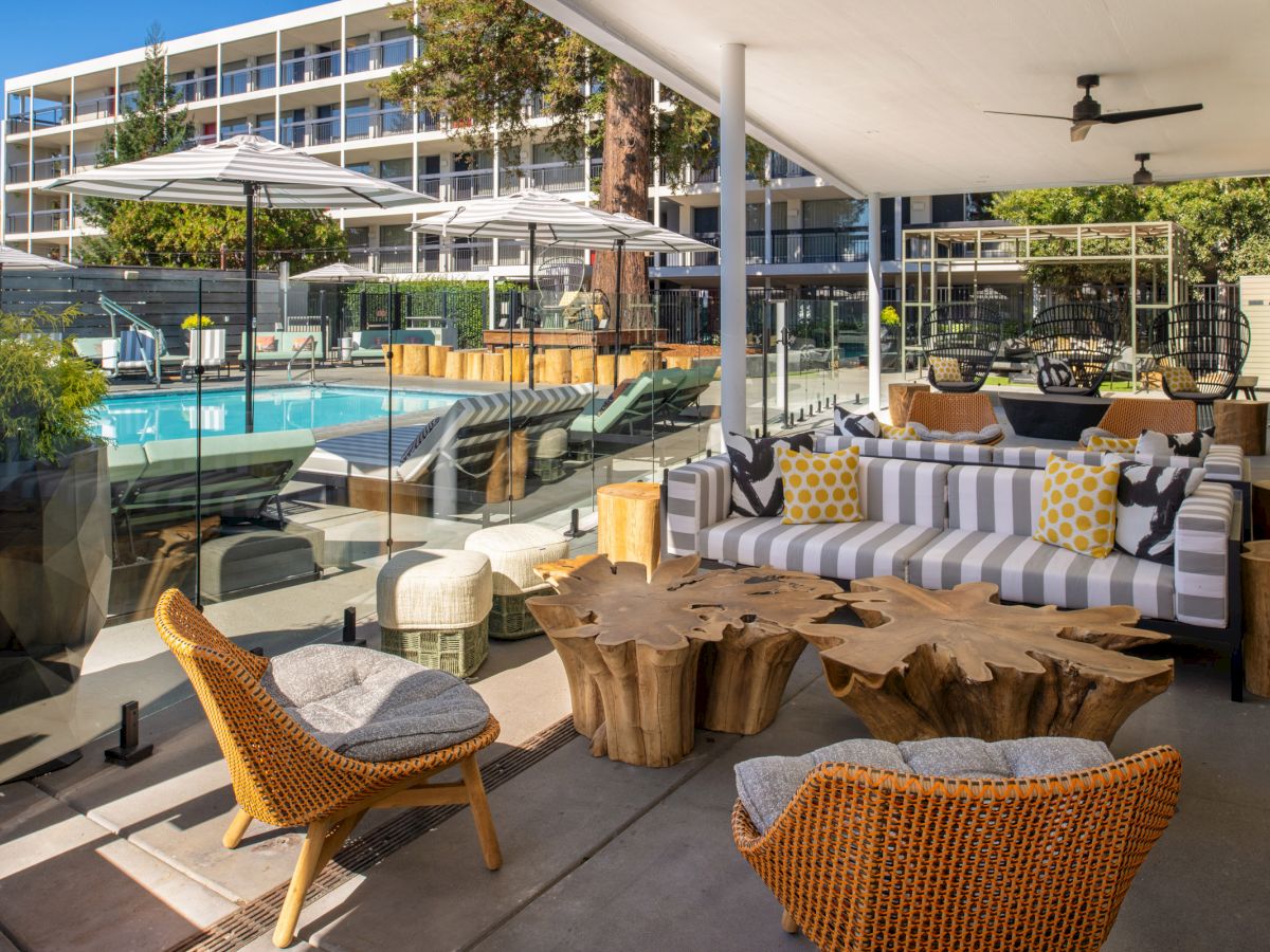 An outdoor lounge area with modern furniture, wooden tables, and a swimming pool in the background, shaded by large umbrellas, all near a multi-story building.