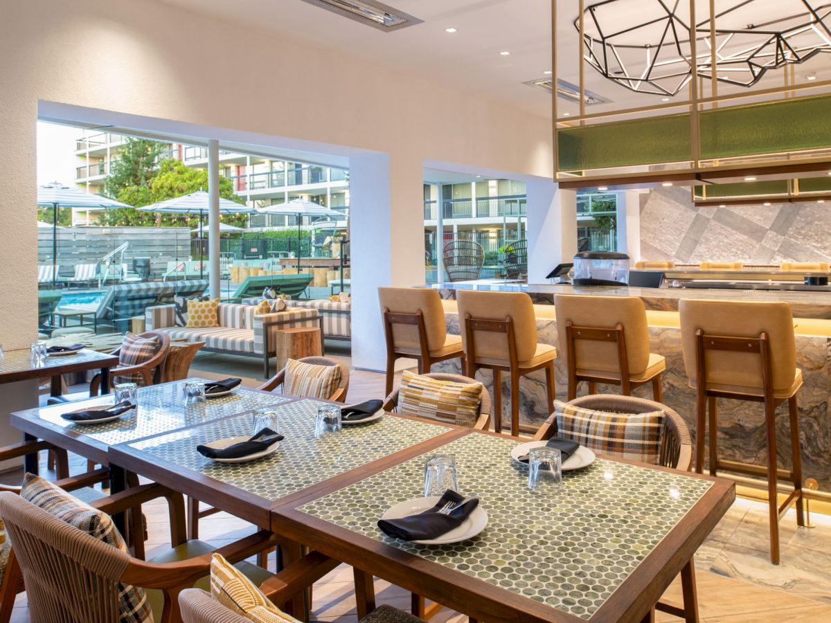 The image shows a modern, well-lit restaurant interior with tables, chairs, a bar area, and large windows offering a view of an outdoor seating area.