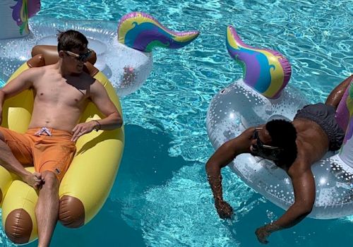 Two men are floating in a pool; one on a yellow float and the other on a unicorn float. Both are enjoying a sunny day.