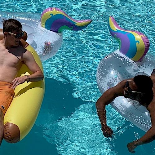 Two men are floating in a pool; one on a yellow float and the other on a unicorn float. Both are enjoying a sunny day.