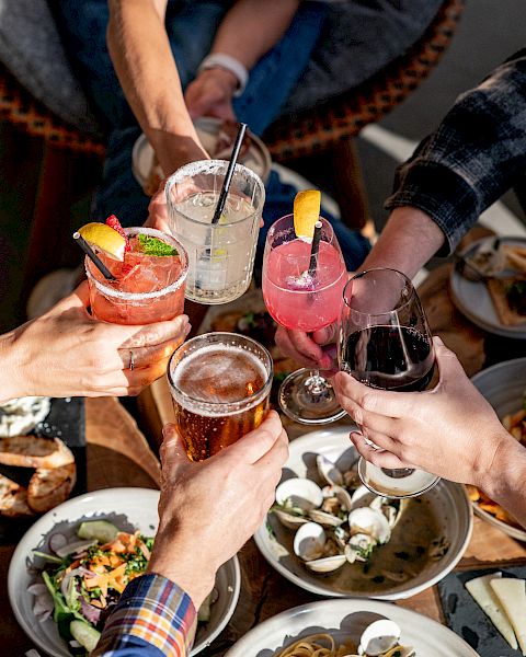 People raising various drinks in a toast, surrounded by plates of food, likely enjoying a social gathering outdoors.