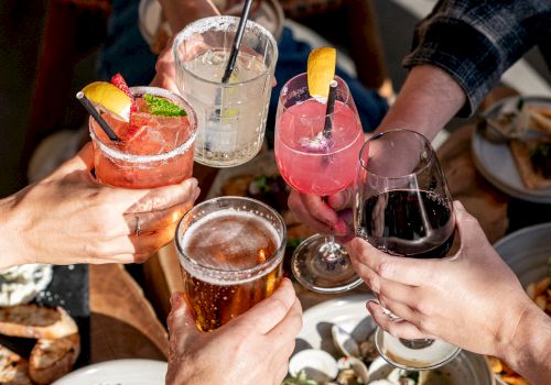 People raising various drinks in a toast, surrounded by plates of food, likely enjoying a social gathering outdoors.