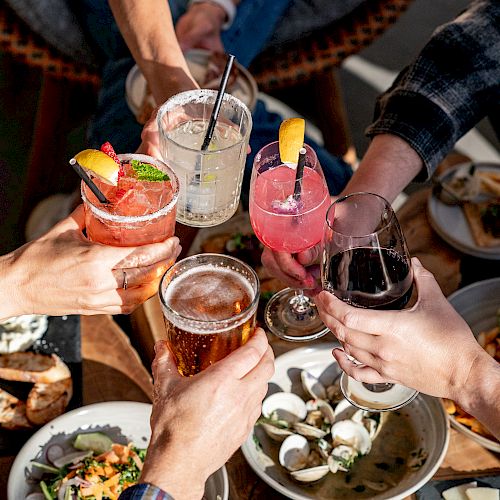 People raising various drinks in a toast, surrounded by plates of food, likely enjoying a social gathering outdoors.