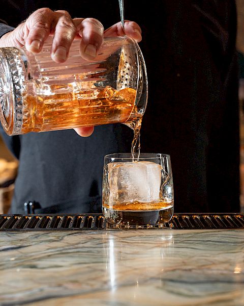 A person is pouring a golden-brown liquid from a glass pitcher into a glass with a large ice cube, placed on a bar counter.