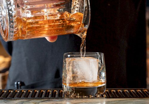 A person is pouring a golden-brown liquid from a glass pitcher into a glass with a large ice cube, placed on a bar counter.