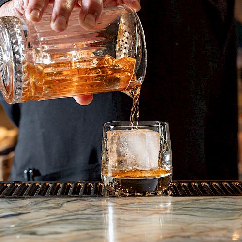 A person is pouring a golden-brown liquid from a glass pitcher into a glass with a large ice cube, placed on a bar counter.