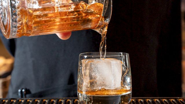 A person is pouring a golden-brown liquid from a glass pitcher into a glass with a large ice cube, placed on a bar counter.